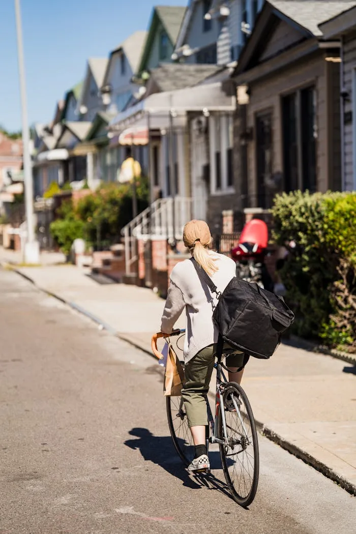 A woman rides a bicycle with a delivery bag on a sunny residential street.